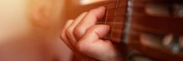 Close-up shot of a hand playing a complex chord on a guitar, with soft focus on the background suggesting a home recording studio. The lighting is warm and focused on the fretboard, highlighting the musician's skill.